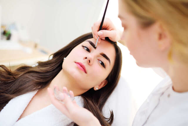 Microblading of eyebrows. A cosmetologist paints the eyebrows of a pretty young woman patient. Semi-permanent makeup for eyebrows.